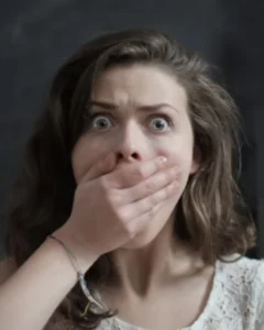 Femme aux yeux grands ouverts, la main sur la bouche, illustrant le silence imposé, les non-dits et l’émotion retenue qui peuvent affecter la santé mentale et l’affirmation de soi.