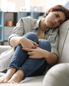 Femme assise sur un canapé, recroquevillée sur elle-même, le regard inquiet et fatigué, illustrant l’anxiété et la vulnérabilité vécues lors d’un retour au travail après un congé de maladie.