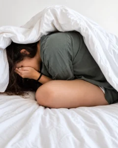 Une femme recroquevillée sur son lit, le visage caché dans ses mains, illustrant l’anxiété et le sentiment de débordement qu’elle peut ressentir au quotidien avec son TDAH.